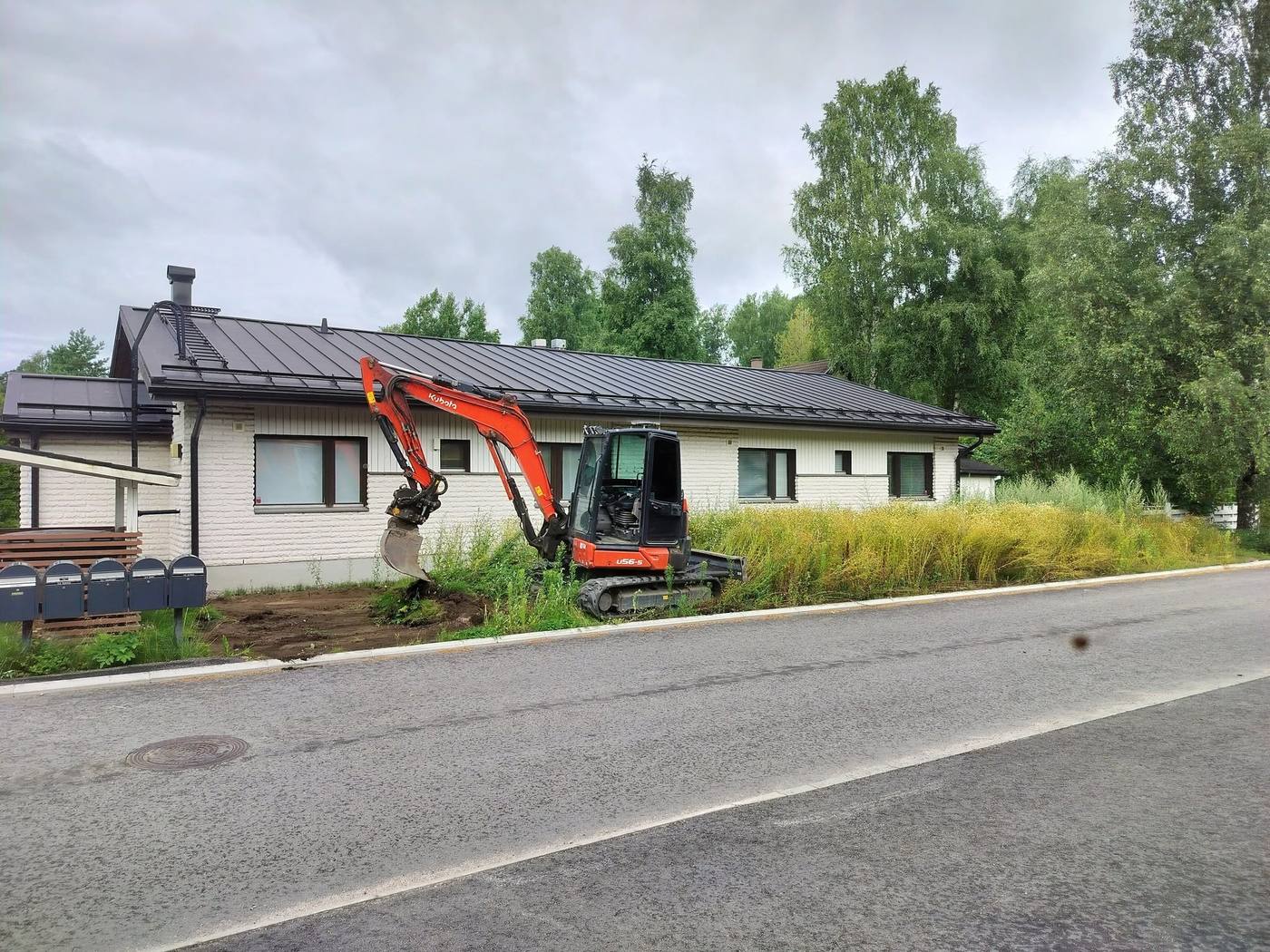 Kaivinkone työskentelee talon edustalla tien varressa, piha auki ja maa möyhennettynä.